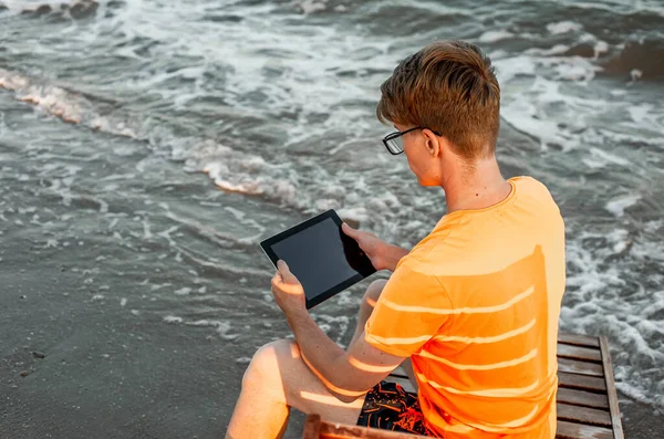 Tişörtlü ve şortlu bir adam tablet tutuyor, deniz kenarındaki ahşap iskemlede oturuyor.