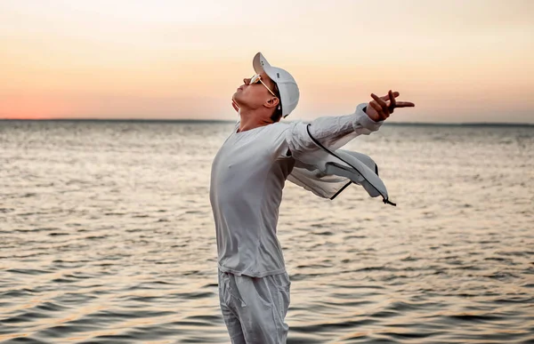 Beyaz elbiseli bir adam deniz kenarında gün batımına bakıyor.