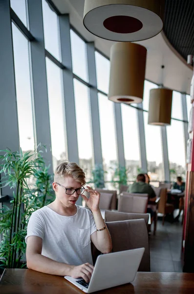 Beyaz tişörtlü ve gözlüklü genç adam kafeteryadaki bir masada dizüstü bilgisayarla çalışıyor.