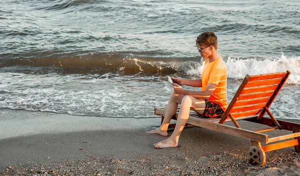 Tişörtlü ve şortlu bir adam tablet tutuyor, deniz kenarındaki ahşap iskemlede oturuyor.