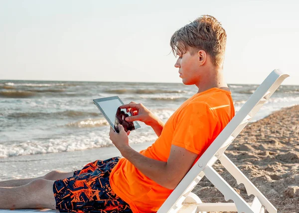 Tişörtlü ve şortlu bir adam tablet tutuyor, deniz kenarındaki ahşap iskemlede oturuyor.