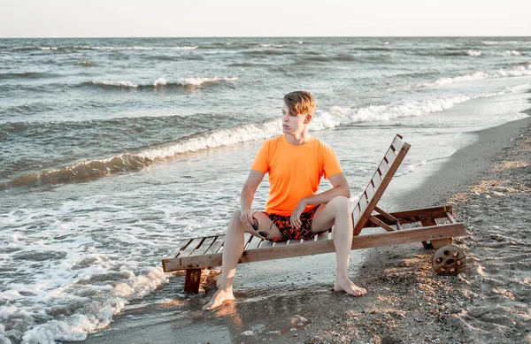 Tişörtlü ve şortlu bir adam deniz kenarındaki ahşap iskemlede oturuyor.