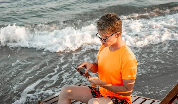 Tişörtlü ve şortlu bir adam tablet tutuyor, deniz kenarındaki ahşap iskemlede oturuyor.