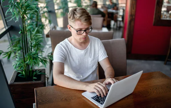 Beyaz tişörtlü ve gözlüklü genç adam kafeteryadaki bir masada dizüstü bilgisayarla çalışıyor.