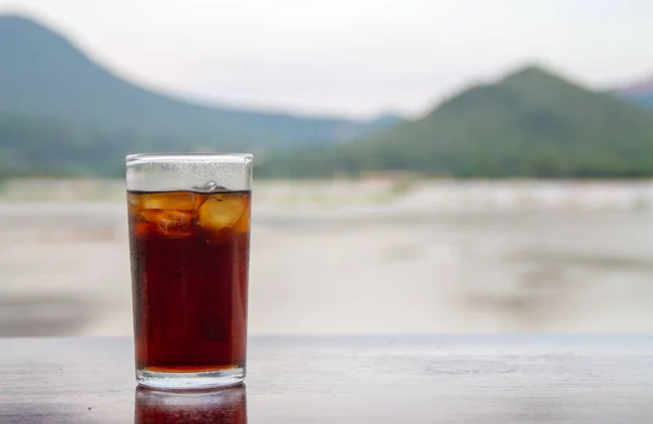 Cola in glass on table with mountain and river background