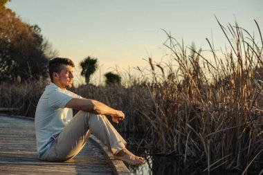 Gün batımında çıplak ayakla gölün kıyısındaki ahşap kaldırımda oturan genç adam. İlham verici bir kavram