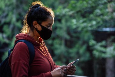 Cep telefonu tutan ve doğal bir ortamda maske takan genç bir adam. Covid-19 salgını ve iletişim konsepti.