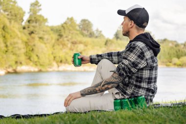 Genç dövmeli adam nehir kenarında bira içiyor çimlerin üzerinde oturuyor..