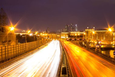 Gece karanlık gökyüzüne karşı farlar ve mimari ışık parlayan hatları ile bir şehir karayolu üzerinde panoramik görünümü