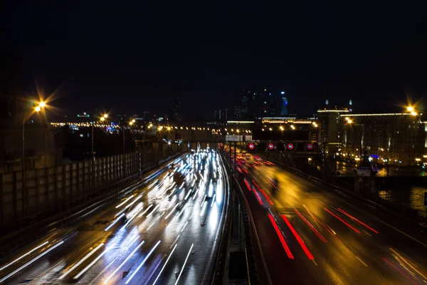 夜の暗い空に対するヘッドランプと建築からの光線の輝く都市高速道路のパノラマビュー — ストック写真