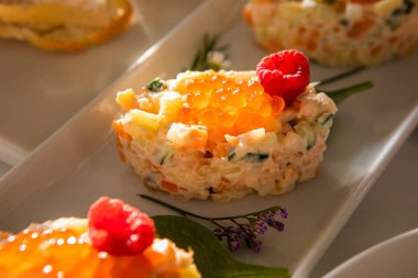 Çiçeklerle süslenmiş bir masada soğuk atıştırmalık. Kırmızı havyarla süslenmiş salata ve beyaz tabakta ahududu. Rus salatası Olivier, bıldırcın yumurtası, havyar, bezelye, et, patates, havuç.
