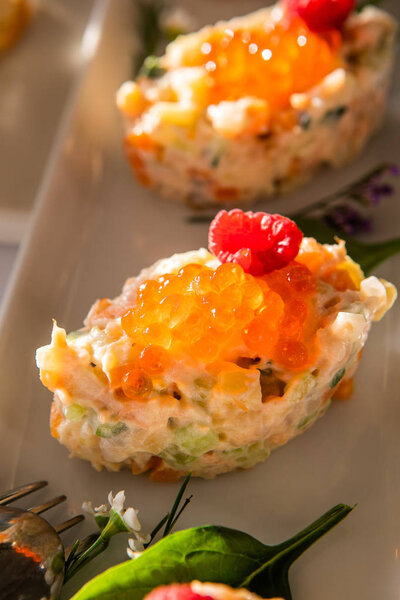 Холодная закуска на праздничном столе, украшенном цветами. Салат украшен красной икрой и малиной на маленькой белой тарелке. Русский салат, Оливье, с перепелиными яйцами, икрой, горохом, мясом, картошкой, морковью
