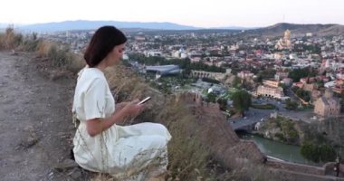 Şehrin yukarısındaki tepede oturan ve cep telefonuyla fotoğraf çeken elbiseli güzel turist kız.