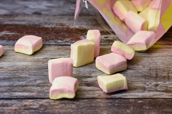 Macro of yellow and pink marshmallows lying in front of a plastic bag on wooden surface — Stock Photo, Image