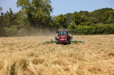 Hay Hasat Haziran 2020 İngiliz kırmızı traktörü, onu baltalamak için saman dönüyor, mavi gökyüzü güneşli bir gün. Güneş parlarken saman yap. Hampshire İngiltere