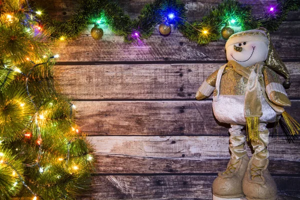 Festive Christmas background of dark old wooden boards, fir-tree, snowman and luminous garland with colored lights and Christmas balls for a banner, postcard, advertisement with place for text
