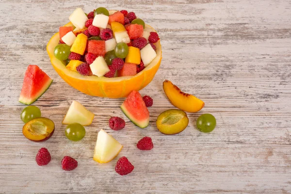 Fruit salad. Peach, watermelon, melon, raspberry, plum, grapes, diced lie in a melon salad bowl on a white wooden background. Pieces of fruit. Place for text. Copy space. Top View, Flat Lay