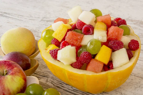 Fruit salad. Peach, watermelon, melon, raspberry, plum, grapes, diced lie in a melon salad bowl on a white wooden background. Whole fruits in wooden spoons. Place for text. Copy space.