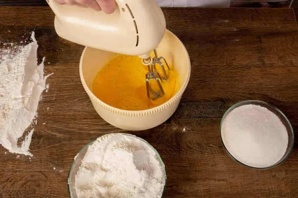 A chef-cook an electric mixer in his hands beats the eggs in a bowl on a wooden table. Ingredients for cooking. Flat Lay, top view, Copy space, place for text.