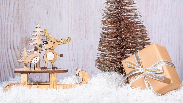 New Year and Christmas background for greetings, banner, postcard, advertisement with place for text. Wooden deer on a sled and a box with a gift close-up on a background of a golden Christmas tree. Copy space, front view.