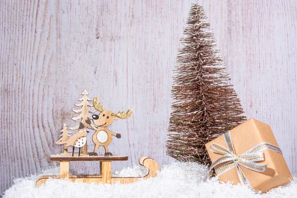 New Year and Christmas background for greetings, banner, postcard, advertisement with place for text. Wooden deer on a sled and a box with a gift close-up on a background of a golden Christmas tree. Copy space, front view.