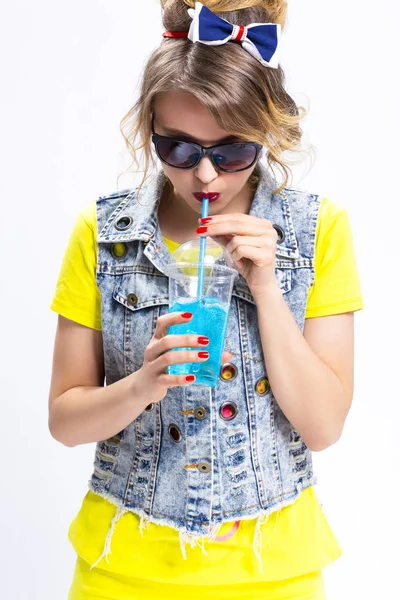 Happy Youth Lifestyle Concepts. Funny and Upbeat Caucasian Blond Girl Drinking Blue Cocktail Through The Straw. Wearing Denim Vest and Sunglasses.Vertical Image Orientation