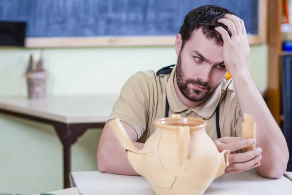 Portrait of Depressed Male Ceramist Sitting With Negative Expression In Front of Broken Jar in Workshop.Staring at Broken Piece. Horizontal image