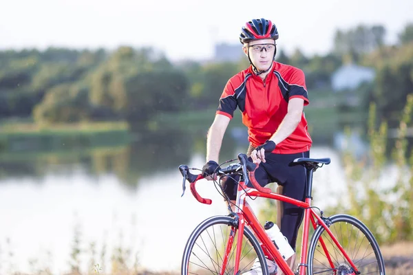 Portrait of Caucasian Male Cyclist in Professional Bike Outfit Posing Outdoors with red Road Bike. Outdoor Shot. Horizontal Image Composition