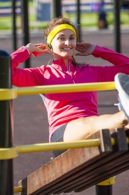 Spor kavramları. Mutlu barfiks uzun bankta yapma açık spor kıyafeti sportif beyaz kız gülümsüyor. Kulaklık müzik dinlemek. Dikey görüntü kompozisyon