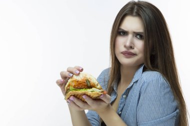 Fastfood kavramı yeme. Genç beyaz kadın yeme hamburger ile nahoş yüz ifadesi Studio. Çizgili gömlek Indoors.Horizontal görüntüde poz