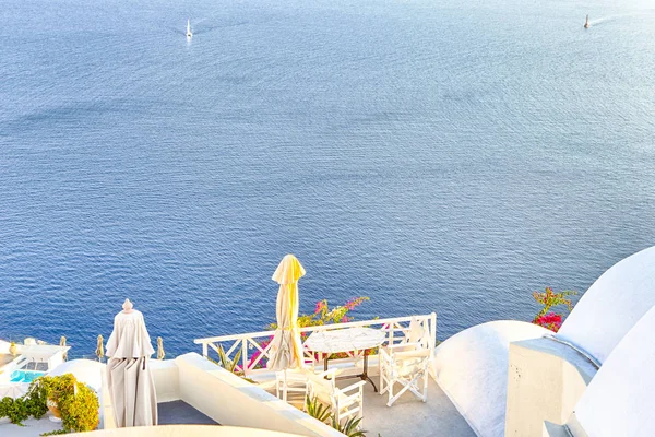 European Scenic Destinations. Pale Houses And Open Air Restaurant of Oia Village in Santorini in Greece with Sailing Boats on Background. Horizontal image