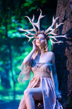 Yaz Ormanında Güzel Geyik Boynuzlarıyla Güzel Kız Poz Veriyor Yüksek Ağacın yanında. Orman Perisi Konsepti için hafif bir elbise giyiyor. Sanat fotoğrafçılığı. Dikey Resim Yönlendirmesi