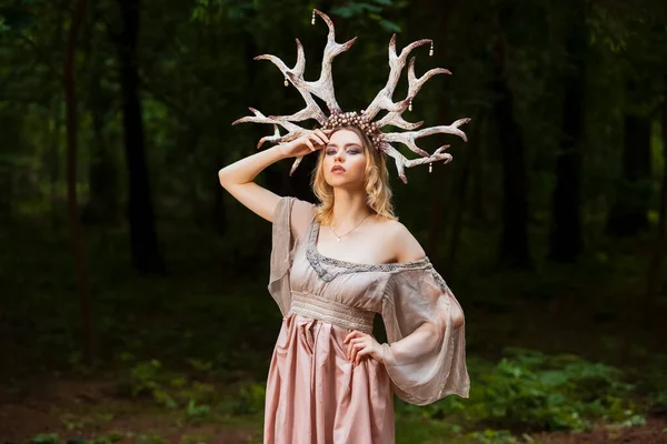 Beautiful Caucasian Girl With Artistic Deer Horns  In Forest. Posing in Light Dress for Demonstrating Forest Nymph Concept for Art Photography. Horizontal Image