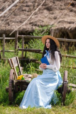 Güzel beyaz tenli esmer kız çiçekli eski moda elbiseler içinde poz veriyor. Köy Çevresindeki Rustik Araba Üzerine Otururken Dışarıda Kitap Okumak. Dikey Resim