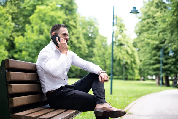 一个年轻的身体适合的商人的肖像在公园里抽烟古巴雪茄和在手机上交谈 — 图库照片