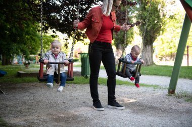 Neşeli Genç Anne ve Bebek Kız ve Erkek İkizler Oyun sahasında Birlikte Oynuyor Onları Salıncakta sallarken Mutlulukla Gülerken