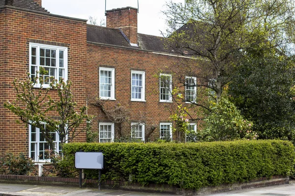 Two-story brick house with white windows. Quiet area in central London, front garden with hedges, spring, flowering trees.