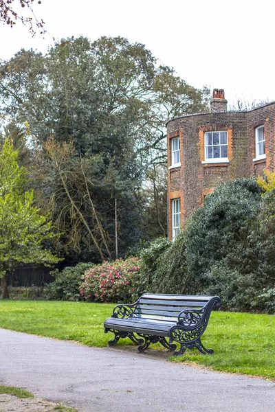 Eine Bank im Park entlang des Weges vor dem Hintergrund eines klassischen englischen Backsteingebäudes — Stockfoto