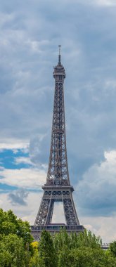 Parlak bulutlu gökyüzünün arka planında ağaçların arkasında Eyfel Kulesi.