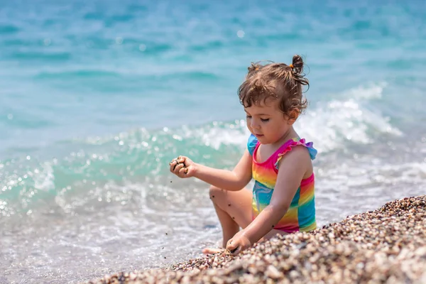 Malá holčička v pestrobarevné plavkách sedí u moře. Holka hraje na pláži. Krásné a zářivě modré moře. — Stock fotografie