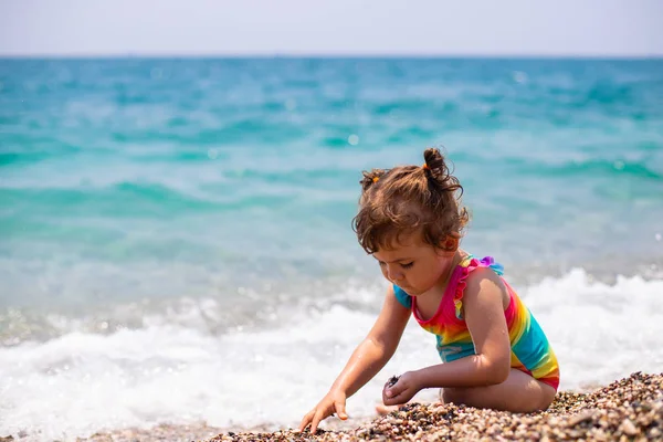 Een klein meisje in een kleurrijk badpak ligt aan de zee. Meisje speelt op het strand. Mooie en heldere blauwe zee. — Stockfoto