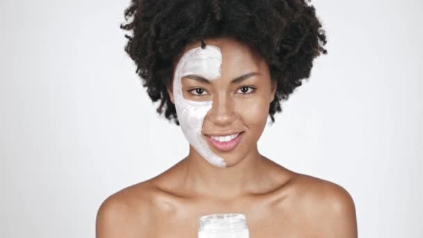 美丽的非洲裔美国女孩在面罩看着容器 采取面霜 涂抹在脸颊上 微笑着在白色 — 图库视频影像