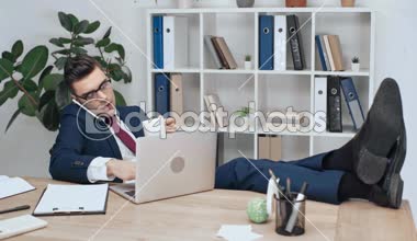 Masa üzerinde çapraz ayaklı işyerinde oturan, smartphone üzerinde konuşmak ve kahve içme laptop kullanma yakışıklı, ciddi iş adamı