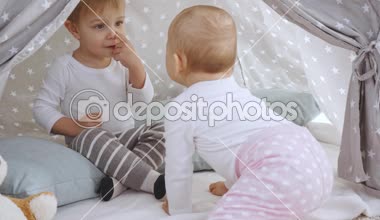 Bebek kız kardeşi tüm fours üzerinde sürünerek ve ona sarılmaya çalışırken bebek Wigwam oturan gülümseyen Toddler çocuk
