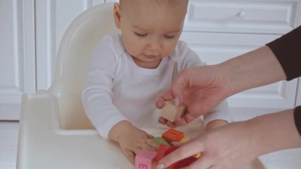 かわいい子供がハイチェアに座って 母は彼女の木製の色とりどりのブロックと赤ちゃんのドロップキューブを与えます — ストック動画