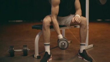 Beyaz şort egzersiz ekipmanları üzerinde oturan sporcu, dumbbell ile eğitim ve pazı bakıyor