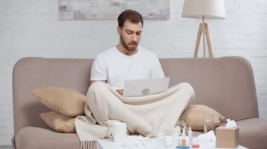 sakallı blogger dizüstü bilgisayar ile kanepede oturan, hapşırma, öksürük, kahve ile fincan alarak ve oturma odasında içme 