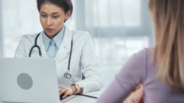 stetoskoplu afro-amerikan doktor hastayla konuşuyor, dizüstü bilgisayar kullanıyor ve klinikte panoya yazı yazıyor