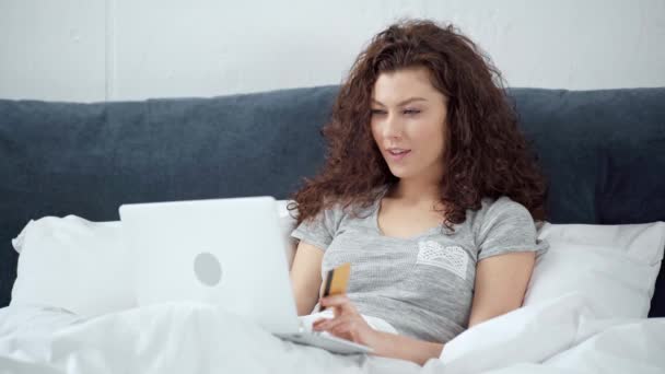 美丽的女孩躺在床上用信用卡 微笑和使用笔记本电脑在家里 — 图库视频影像