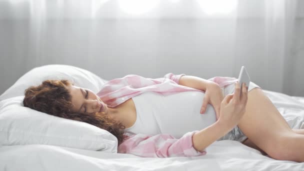 妊娠中の若い女性がベッドに横たわり 笑顔でスマートフォンで自分撮りをする — ストック動画